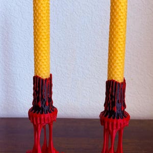 May include: Two yellow beeswax candles with a honeycomb texture, set in red and black candle holders. The holders have a dripping wax design, creating a unique and decorative look. The candles are a warm, inviting color.