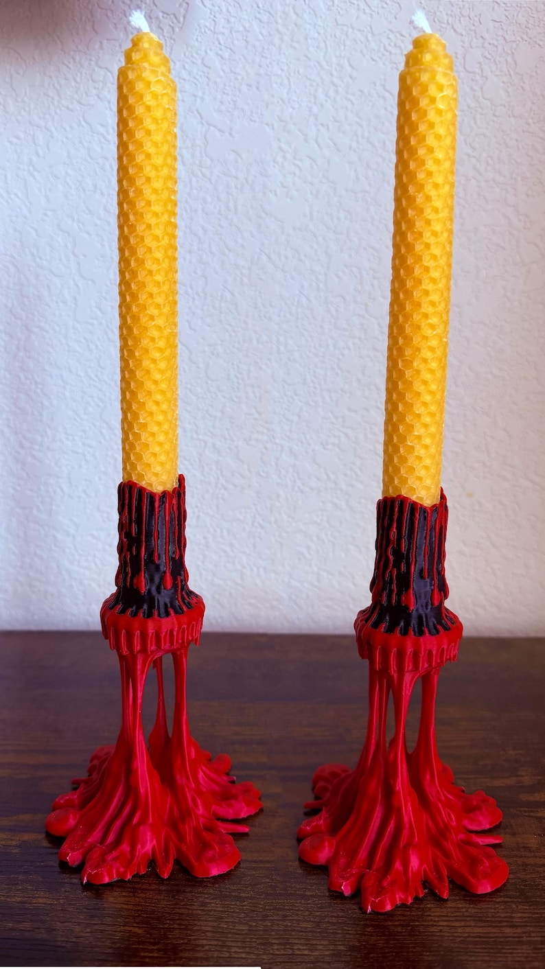 May include: Two yellow beeswax candles with a honeycomb texture, set in red and black candle holders. The holders have a dripping wax design, creating a unique and decorative look. The candles are a warm, inviting color.