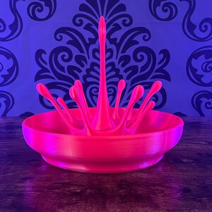 May include: A vibrant pink, 3D-printed splash-shaped bowl. The bowl has a wide, shallow base with a central, upward-reaching splash design. The background features a blue wall with a dark floral pattern.