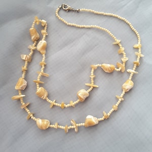 May include: A long, cream-colored necklace with a silver clasp. The necklace is made of small, round beads and larger, irregular-shaped beads that are a slightly darker shade of cream.