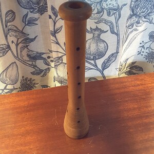 May include: A wooden spindle with a cylindrical body and a flared base. The spindle has a circular top and several small holes along its length. The wood has a light brown color and is set on a wooden surface.