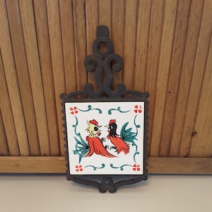 May include: A black cast iron hot pad with a white ceramic tile featuring a colorful illustration of two roosters. The tile has a green, white, and red floral border.