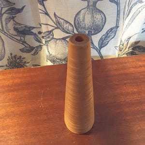 May include: A wooden spool with a conical shape, featuring a hollow center. The spool is a light brown color and sits on a dark brown wooden surface. The background includes a patterned fabric with blue and white botanical designs.