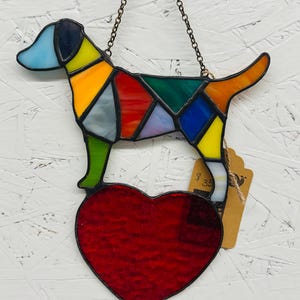 May include: A stained glass dog with a colorful patchwork design, hanging from a chain. The dog is perched above a red heart. The dog's head is light blue, and the body features yellow, orange, green, and blue sections.