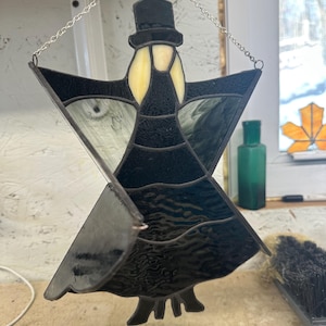 Puede incluir: Adorno de ángel de vidriera con sombrero de copa y vestido negros. La cara del ángel es de color amarillo pálido. Las alas son una mezcla de vidrio transparente y negro. El adorno cuelga de una cadena plateada.