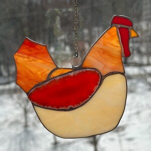 Op de afbeelding: Een gebrandschilderde glazen haan-zonnevanger met een ketting om op te hangen. De haan heeft een rode kam en lellen, een oranje en gele body en een cr&egrave;mekleurige buik. Het glas heeft een getextureerd uiterlijk.