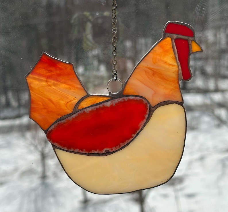 Op de afbeelding: Een gebrandschilderde glazen haan-zonnevanger met een ketting om op te hangen. De haan heeft een rode kam en lellen, een oranje en gele body en een cr&egrave;mekleurige buik. Het glas heeft een getextureerd uiterlijk.