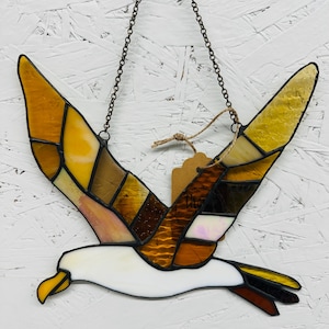 Peut inclure: Vitrail représentant une mouette en vol, composé de pièces de verre ambré, marron et blanc. L'oiseau a les ailes déployées et un bec jaune, suspendu par une chaîne. Une petite étiquette est suspendue.