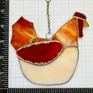 Op de afbeelding: Gebrandschilderd glazen haan zonnevanger met een ketting om op te hangen. De haan heeft een rode kam en lel, met oranje, geel en wit glas. De body is ongeveer 13 cm hoog.