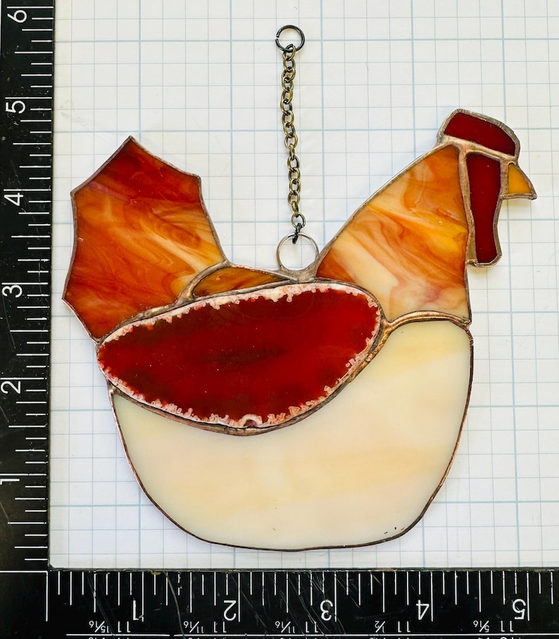 Op de afbeelding: Gebrandschilderd glazen haan zonnevanger met een ketting om op te hangen. De haan heeft een rode kam en lel, met oranje, geel en wit glas. De body is ongeveer 13 cm hoog.