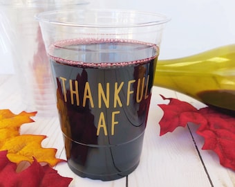 May include: A clear plastic disposable cup filled with dark red liquid, featuring the gold text THANKFUL AF printed on the front. The cup is positioned among several artificial autumn leaves in shades of red, orange, and yellow.