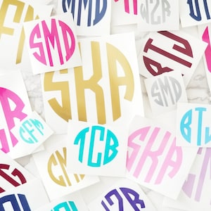 May include: A collection of circular vinyl decals featuring various three-letter monograms in diverse colors including gold, hot pink, cyan, navy blue, burgundy, light pink, and lavender, arranged in a scattered pattern on a white surface.