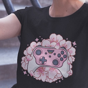 Peut inclure: T-shirt noir avec un motif de manette de jeu rose et blanche entourée de fleurs de cerisier roses et d'étoiles. La manette comporte une croix directionnelle et plusieurs boutons. Le motif est centré sur le devant du t-shirt.
