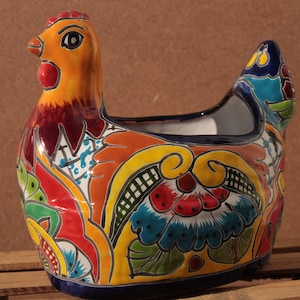 Op de afbeelding: Een kleurrijke keramische kippenpot met een blauw, geel, rood, groen en wit bloemenontwerp. De pot heeft een brede opening om te planten.