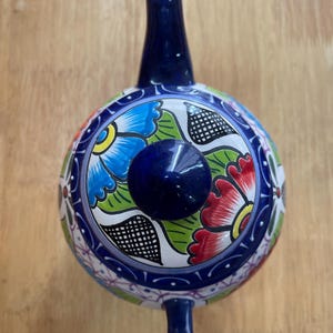 May include: A blue and white ceramic teapot with a floral design. The teapot has a round body and a spout on the side. The lid is decorated with a large flower in shades of red, blue, green, and yellow.