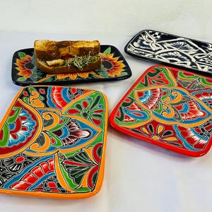 May include: A collection of colorful, hand-painted ceramic serving platters. One platter holds a sandwich. The platters feature vibrant floral and abstract designs in red, orange, blue, green, and black. The platters are rectangular and square-shaped.