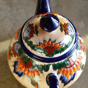 May include: A white ceramic teapot with a blue handle and lid. The teapot is decorated with colorful floral designs, including sunflowers, in shades of orange, red, blue, and green.
