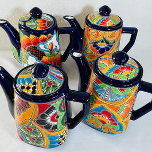 May include: Four ceramic teapots featuring vibrant, hand-painted floral and geometric patterns in shades of blue, orange, red, green, and yellow. Each teapot has a dark blue handle, spout, and lid with a matching knob, showcasing intricate designs.