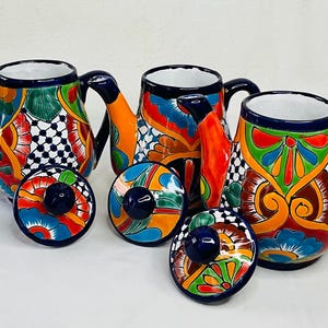 Puede incluir: Tres teteras de cerámica coloridas con tapas, que presentan intrincados patrones florales y geométricos en tonos de azul, verde, naranja y amarillo. Las teteras tienen un estilo tradicional mexicano Talavera.