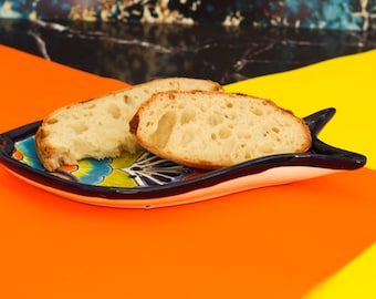 Puede incluir: Dos rebanadas de pan crujiente en un plato de cerámica en forma de pez azul y blanco. El plato está sobre una superficie naranja brillante.