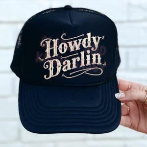 May include: Black trucker hat with a gold embroidered design that says "Howdy Darlin'" on the front.