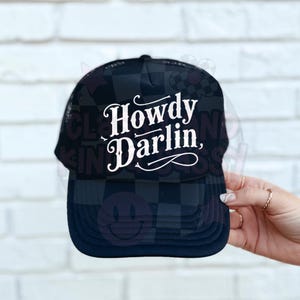 May include: Black and navy blue trucker hat with a checkered pattern on the front and brim. The hat features the text "Howdy Darlin'" in white script across the front.