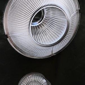 May include: Two clear glass objects with a textured, ridged design. The larger object is circular with a central opening and a detailed, patterned surface. The smaller object is a circular plate with a radial pattern. Both objects are against a dark background.