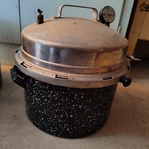May include: A vintage pressure cooker with a speckled black and white enamel pot and a silver-colored lid. The lid has a handle, a pressure gauge, and a vent. The cooker is designed for cooking food under pressure.