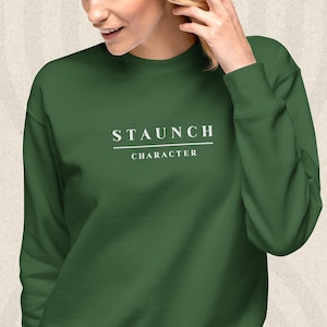 May include: A forest green crewneck sweatshirt featuring the white text STAUNCH CHARACTER printed on the chest. The design includes a horizontal line separating the two words, with the word STAUNCH above the line and CHARACTER below it.