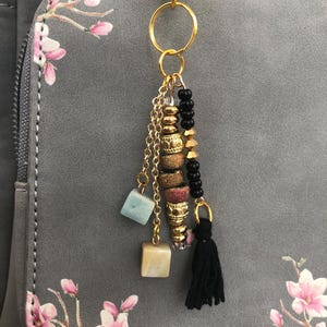 May include: A gold-toned metal keychain featuring a central ring with multiple dangling elements, including a black fabric tassel, a strand of black beads, a strand of textured gold-toned beads, and two small square-shaped stone charms in blue and cream.