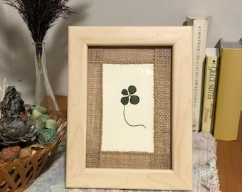 May include: A framed artwork featuring a four-leaf clover illustration. The artwork is set against a burlap background within a light wood frame. The clover is depicted in dark green on a white paper. The frame is displayed on a patterned surface.