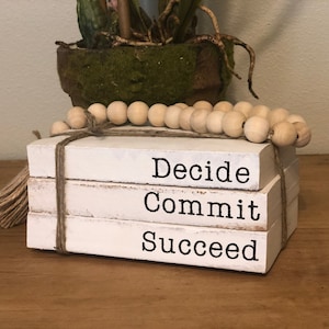 May include: A stack of three white faux books with the words "Decide", "Commit", and "Succeed" printed in black. The books are tied together with twine and decorated with a strand of wooden beads.