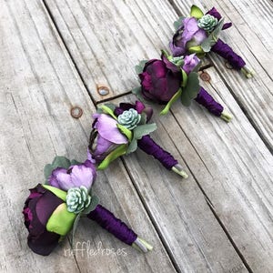 Könnte beinhalten: Vier violette und grüne Ansteckblumen mit Sukkulenten und einem violetten Band. Die Ansteckblumen sind in einer Reihe auf einer Holzoberfläche angeordnet.