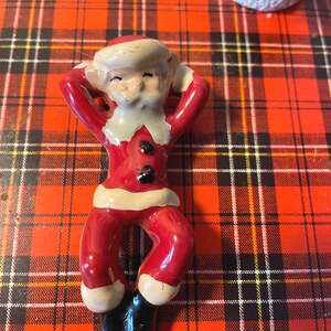 May include: A ceramic figurine of Santa Claus in a red and white suit, with his arms behind his head, relaxing on a red and black plaid surface.