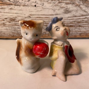 May include: A vintage porcelain figurine set of a cat holding a red apple and a person with blue hair wearing a yellow and red outfit.