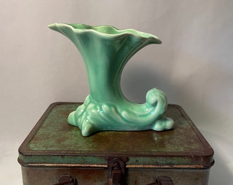 May include: A light green ceramic vase with a unique, curved design. The vase has a wide, flared opening and a long, curved spout. It is sitting on a vintage metal box with a rusty patina.
