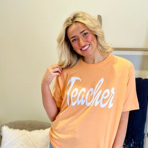 May include: A woman wearing a peach colored t-shirt with the word "Teacher" in white cursive lettering. She is wearing blue jeans with holes in the knees.