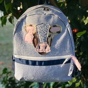 May include: A blue and white striped backpack with a cow applique. The cow has brown and pink features and a floral pattern on its head.