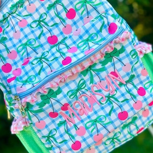 May include: A light blue and white gingham backpack featuring a pattern of pink and red cherries with green bows. The front pocket has a ruffled trim and the name MADISYN embroidered in pink thread. The sides are accented with green polka dot fabric.