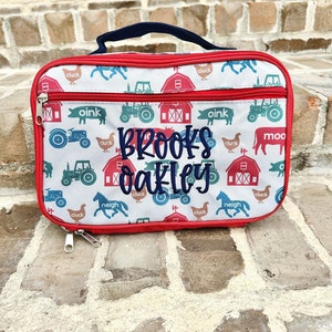 May include: Red and white lunch bag with a farm animal theme. The bag has a zipper closure and a navy blue handle. The bag is personalized with the name "Brooks Oakley" in blue embroidery.