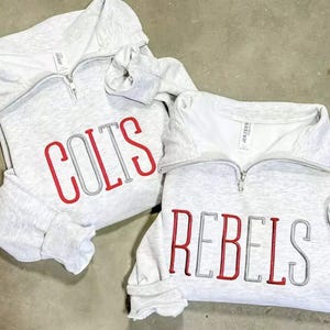 May include: Two light gray sweatshirts with quarter-zip collars. One sweatshirt has the word "COLTS" embroidered in red and gray. The other sweatshirt has the word "REBELS" embroidered in red and gray.