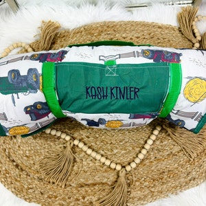 May include: A rolled-up, personalized play mat featuring a farm theme. The mat has a white background with illustrations of tractors, hay bales, and fences. A green panel with the name "KOSH KINLER" is embroidered on the front. The mat has a green handle.