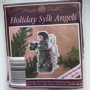 May include: A Mill Hill Holiday Sylk Angels craft kit featuring a silver moon angel with a green dress and a red ribbon. The kit includes glass beads, perforated paper, floss, needles, a pin back, and instructions. The kit is 36 inches by 25 inches.  MHAS12 Silver Moon Angel. 1995, Mill Hill.