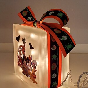 May include: A glass block with a Halloween-themed image of Mickey Mouse, Minnie Mouse, Pluto, and Goofy dressed in costumes. The block is lit with white fairy lights and wrapped with an orange and black ribbon with a skull and crossbones pattern.