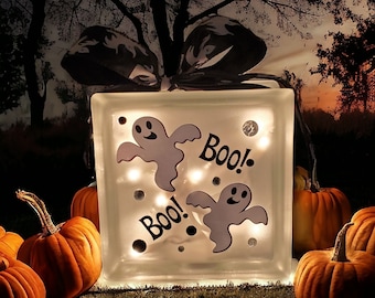 May include: A white glass block with a frosted finish features two ghost silhouettes and the text "Boo!" repeated twice. The block is illuminated from within, creating a warm glow.  The block is surrounded by pumpkins.