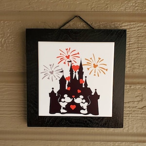 May include: A black framed tile with a white background featuring a silhouette of Cinderella's castle with Mickey and Minnie Mouse in front of it.  The castle has red hearts on the towers and there are fireworks in the background with red, orange, and gray hearts.