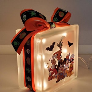 May include: A frosted glass block with a Halloween-themed image of Mickey Mouse, Minnie Mouse, Donald Duck, Goofy, and Pluto dressed in costumes. The block is lit with white fairy lights and decorated with an orange and black ribbon with skulls and pumpkins.