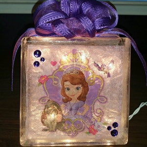May include: A glass block light with a purple ribbon and a princess character image. The image features a princess in a purple dress with a crown and a small animal beside her. The block has purple glitter and rhinestones.