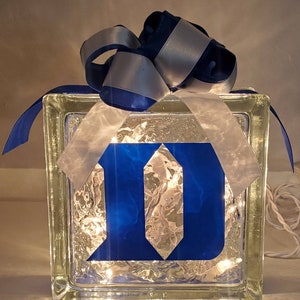May include: A glass block with a blue "D" inside, illuminated with white lights. The block is wrapped with a white ribbon and a blue and silver bow.
