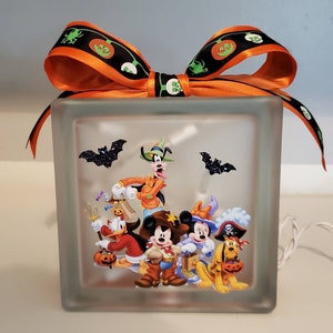 May include: A clear glass block with a Halloween-themed image of Mickey Mouse, Minnie Mouse, Goofy, Donald Duck, and Pluto dressed in costumes. The block is lit from within and has a black and orange ribbon tied around the top.